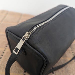 May include: A black rectangular bag with a silver zipper closure. The bag has a small handle and a long strap. The material appears to be leather or a leather-like material. The bag is shown on a patterned surface.
