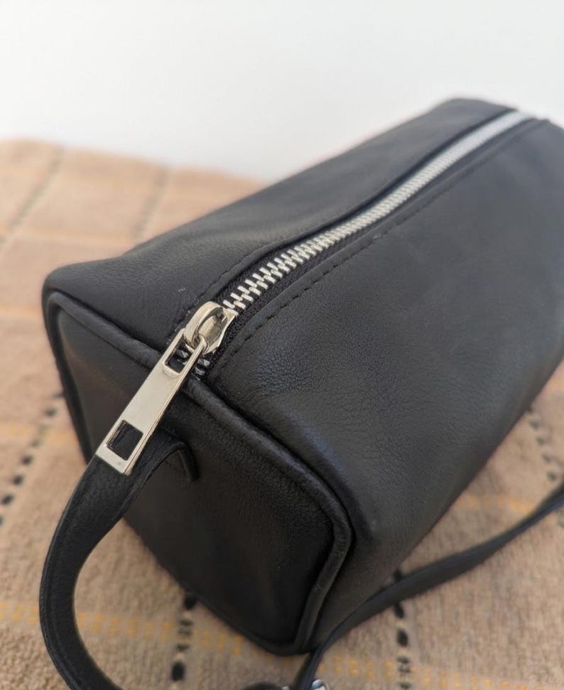 May include: A black rectangular bag with a silver zipper closure. The bag has a small handle and a long strap. The material appears to be leather or a leather-like material. The bag is shown on a patterned surface.