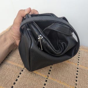 May include: A black leather handbag with a top handle and a silver zipper. The bag is held open, revealing the interior. The leather has a textured appearance. The bag is sitting on a brown surface.