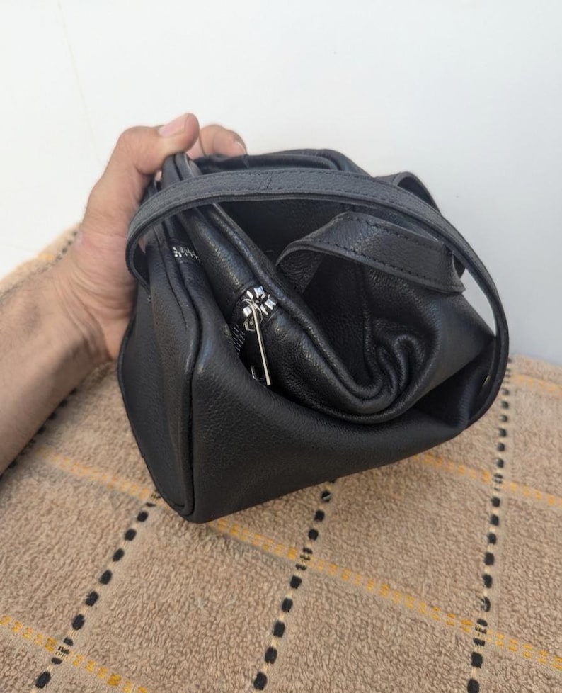 May include: A black leather handbag with a top handle and a silver zipper. The bag is held open, revealing the interior. The leather has a textured appearance. The bag is sitting on a brown surface.