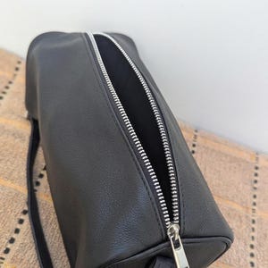 May include: Black cylindrical bag with a silver zipper. The bag is made of a smooth, matte material and has a small strap. The zipper is open, revealing the interior of the bag. The bag is sitting on a patterned surface.