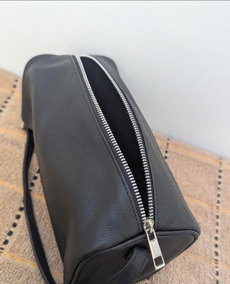 May include: Black cylindrical bag with a silver zipper. The bag is made of a smooth, matte material and has a small strap. The zipper is open, revealing the interior of the bag. The bag is sitting on a patterned surface.
