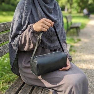 May include: A black rectangular crossbody bag with a silver zipper and a thin shoulder strap. The bag is held by a person sitting on a wooden bench. The background is a blurred outdoor scene with trees and a pathway.