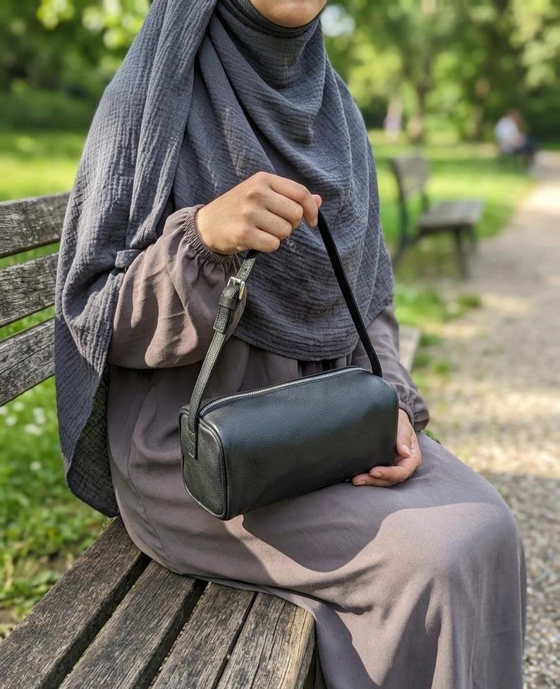 May include: A black rectangular crossbody bag with a silver zipper and a thin shoulder strap. The bag is held by a person sitting on a wooden bench. The background is a blurred outdoor scene with trees and a pathway.
