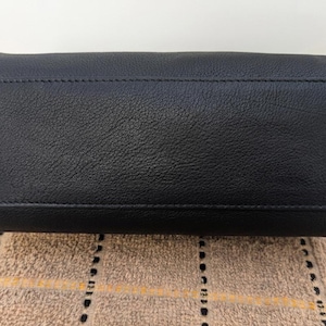 May include: A black leather handbag with a textured surface. The bottom of the bag is rectangular with a stitched seam running across the top. The bag is sitting on a patterned tan surface.