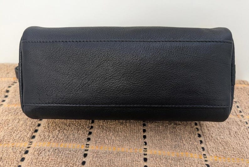 May include: A black leather handbag with a textured surface. The bottom of the bag is rectangular with a stitched seam running across the top. The bag is sitting on a patterned tan surface.