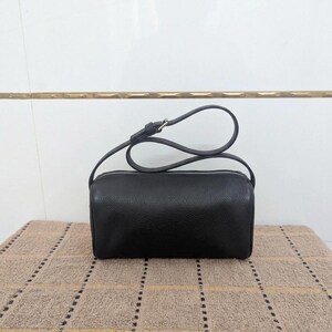 May include: A black, cylindrical crossbody bag with a silver zipper and a long, thin strap. The bag is made of a textured material and sits on a patterned surface.