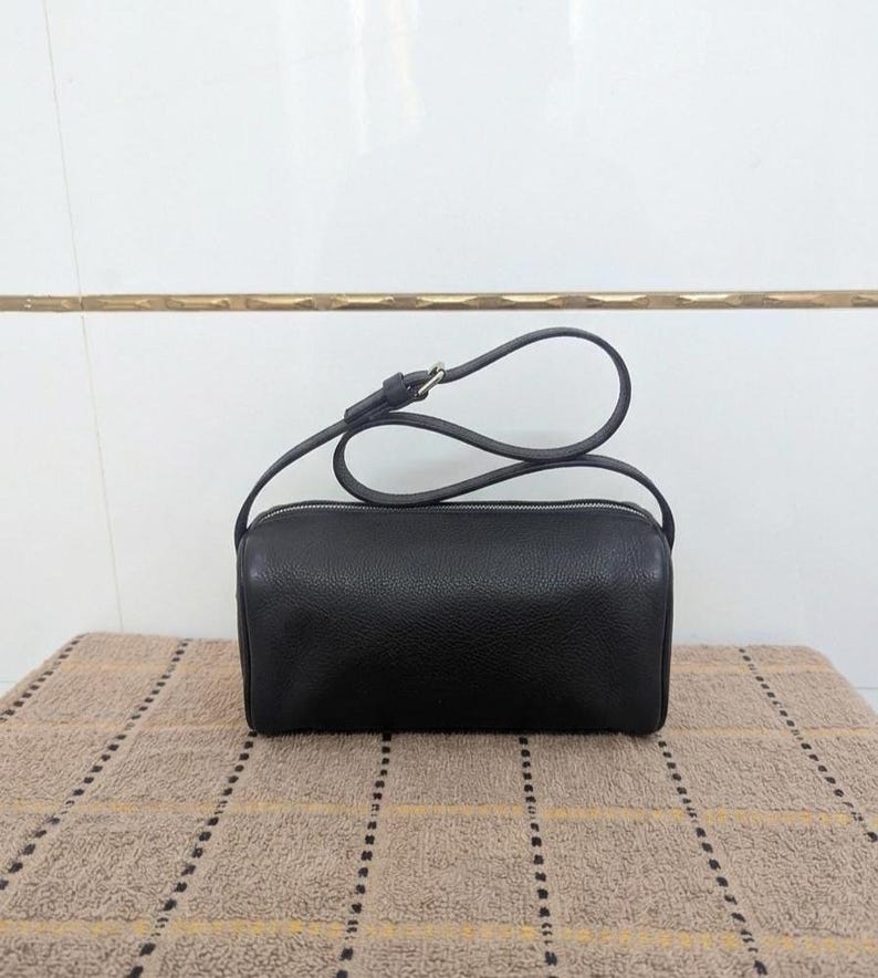 May include: A black, cylindrical crossbody bag with a silver zipper and a long, thin strap. The bag is made of a textured material and sits on a patterned surface.