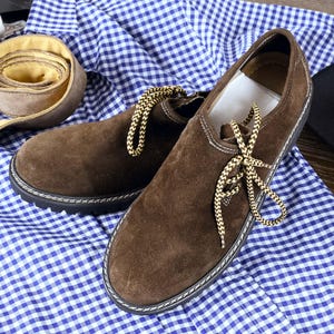 May include: A pair of dark brown suede shoes featuring a rounded toe, a black lug sole with white contrast stitching, and yellow and brown braided laces. The shoes are placed on a blue and white gingham patterned fabric next to a rolled belt.