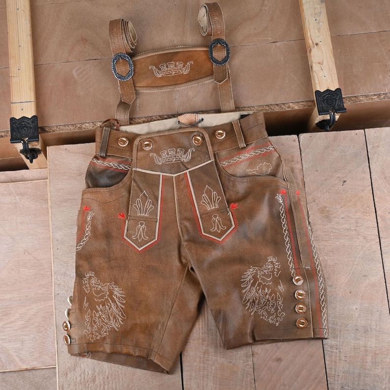 May include: Brown leather lederhosen shorts featuring adjustable suspenders with metal buckles, a front drop-front flap, and intricate white and red embroidery. The garment includes side lacing details, metal buttons along the legs, and a decorative chest bib.