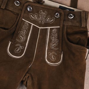 May include: Dark brown suede leather lederhosen featuring a front drop-front flap with cream-colored embroidery of oak leaves and acorns. The garment includes silver-toned metal buttons along the waistband and the central flap.