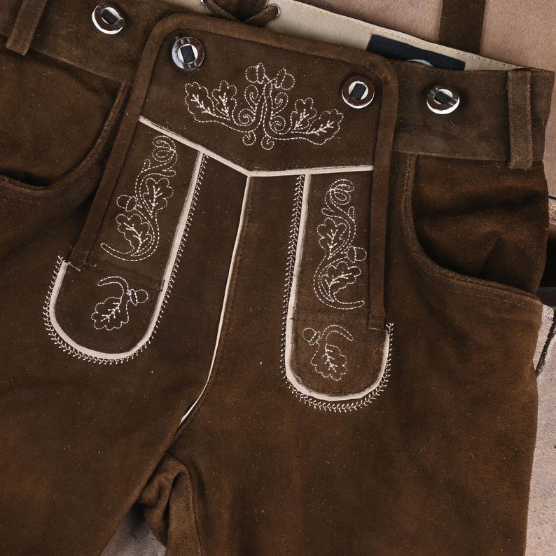 May include: Dark brown suede leather lederhosen featuring a front drop-front flap with cream-colored embroidery of oak leaves and acorns. The garment includes silver-toned metal buttons along the waistband and the central flap.