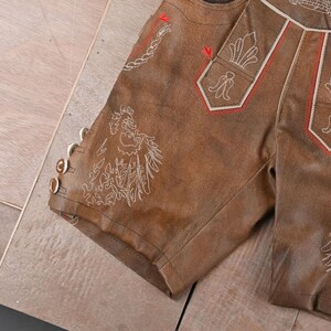 May include: Brown leather shorts featuring intricate white embroidery of a bird and floral motifs, accented with red stitching details. The side of the leg includes three small, round, light-colored buttons for closure and decorative detail.