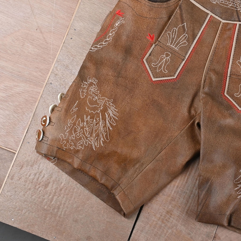 May include: Brown leather shorts featuring intricate white embroidery of a bird and floral motifs, accented with red stitching details. The side of the leg includes three small, round, light-colored buttons for closure and decorative detail.