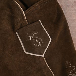 May include: A dark brown suede garment featuring a pentagonal pocket with white stitched embroidery of an acorn and oak leaf. A partial white stitched outline of a deer is visible at the bottom right corner, with light-colored piping along the seams.