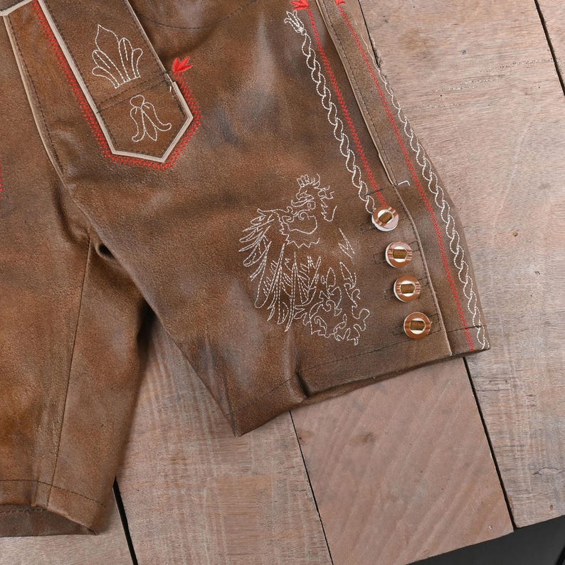 May include: Brown leather lederhosen shorts featuring intricate white embroidery of a bird and floral motifs, accented with red stitching along the seams and four metallic buttons on the side leg panel, showcasing a textured finish.