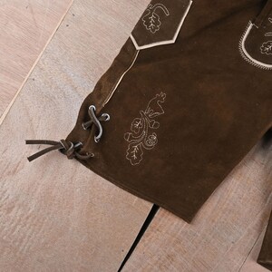 May include: Dark brown suede leather shorts featuring light-colored decorative embroidery of leaves and a deer. The side of the leg includes a lace-up detail with brown leather cords threaded through metal eyelets and a pocket with light piping.