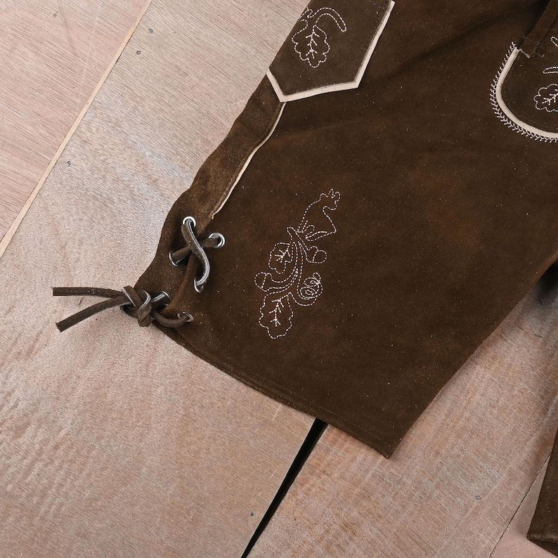 May include: Dark brown suede leather shorts featuring light-colored decorative embroidery of leaves and a deer. The side of the leg includes a lace-up detail with brown leather cords threaded through metal eyelets and a pocket with light piping.