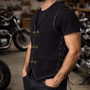 Side angle view of a black suede leather biker vest worn by a male model, showcasing adjustable side lacing with metal eyelets for a customizable fit. The vest features triple front hook closures, braided trim edges, and a fitted silhouette. The model stands slightly turned in a motorcycle workshop setting, emphasizing the vest’s biker aesthetic and breathable sleeveless design.