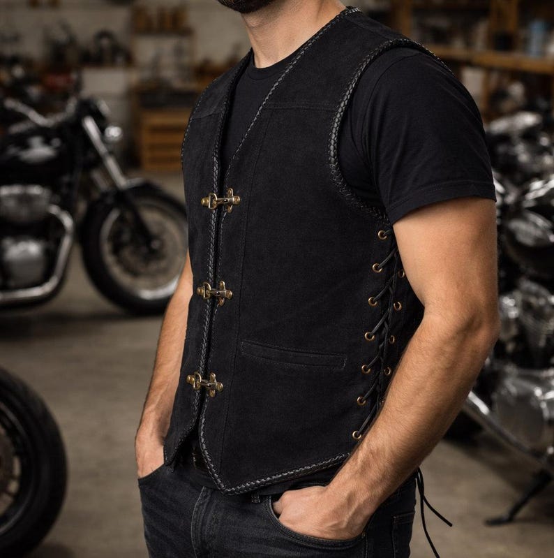 Side angle view of a black suede leather biker vest worn by a male model, showcasing adjustable side lacing with metal eyelets for a customizable fit. The vest features triple front hook closures, braided trim edges, and a fitted silhouette. The model stands slightly turned in a motorcycle workshop setting, emphasizing the vest’s biker aesthetic and breathable sleeveless design.