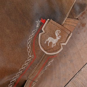 May include: A brown suede leather garment featuring a shield-shaped patch with an embroidered white stag. The surface is decorated with intricate white rope-style stitching and parallel red zigzag embroidery lines that follow the seam.