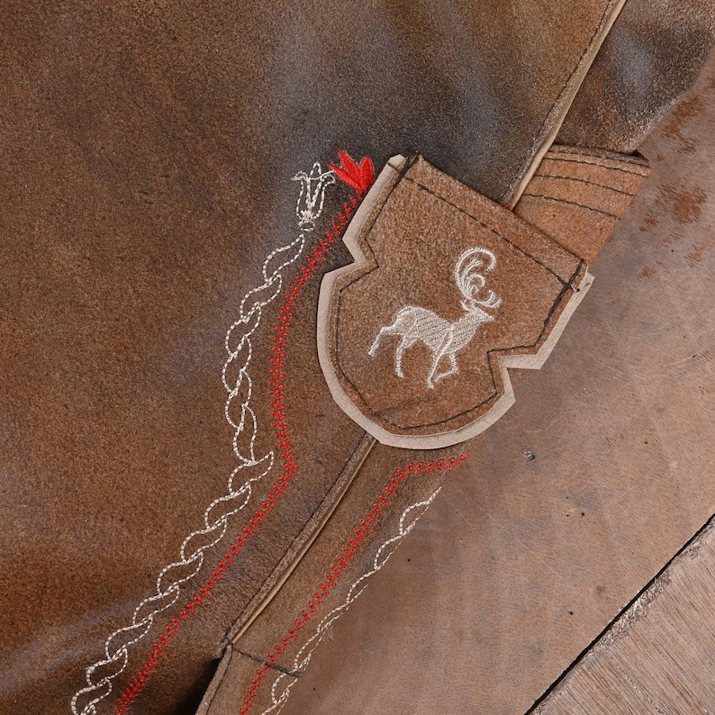May include: A brown suede leather garment featuring a shield-shaped patch with an embroidered white stag. The surface is decorated with intricate white rope-style stitching and parallel red zigzag embroidery lines that follow the seam.