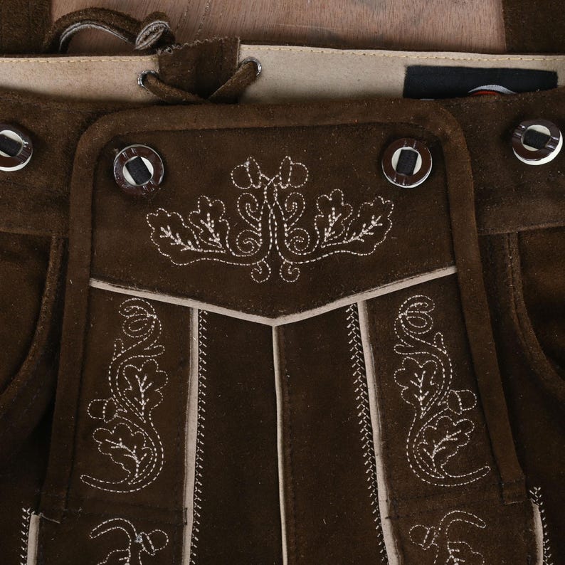 May include: Dark brown suede leather lederhosen featuring light-colored decorative embroidery of oak leaves and acorns. The garment includes a front flap with four dark buttons, a lace-up detail at the waistband, and light-colored piping accents.