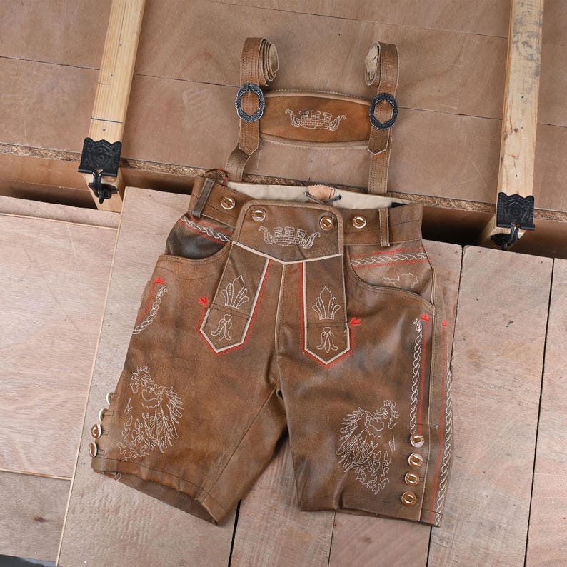 Brown leather lederhosen shorts with suspenders. The shorts feature decorative embroidery in white and red, with ornate designs on the pockets and sides. The suspenders have metal buckles and are attached to the shorts.