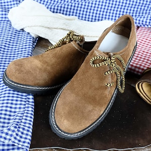Pode incluir: Um par de sapatos de camurça castanha com biqueira arredondada, sola grossa de borracha preta com costuras brancas em contraste e atacadores com padrão amarelo e preto. Os sapatos têm um design de cano baixo com acabamento suave.