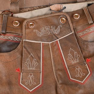 May include: Brown leather lederhosen featuring a front drop-front flap with white embroidered crown and floral motifs. The garment includes red decorative stitching along the edges, belt loops, and metallic buttons on the waistband.