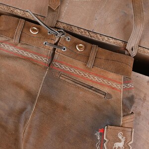 May include: Brown leather breeches featuring a lace-up front closure with metal eyelets, belt loops, and suspender straps. The garment is detailed with red embroidered rope-style patterns and a white embroidered deer motif on a side pocket.