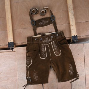 May include: Dark brown leather lederhosen featuring light-colored embroidery of floral and leaf motifs on the front flap, pockets, and legs. The shorts include adjustable suspenders with metal buckles and lace-up details at the leg hems.