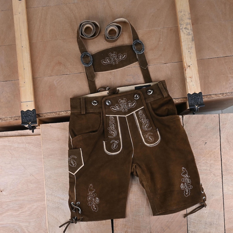 May include: Dark brown leather lederhosen featuring light-colored embroidery of floral and leaf motifs on the front flap, pockets, and legs. The shorts include adjustable suspenders with metal buckles and lace-up details at the leg hems.