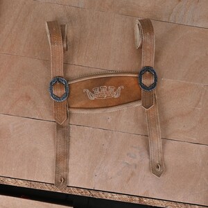 May include: A pair of tan leather suspenders featuring a central horizontal chest strap with an embroidered white castle emblem. The suspenders include two adjustable vertical straps secured by dark metal oval buckles with intricate embossed detailing.