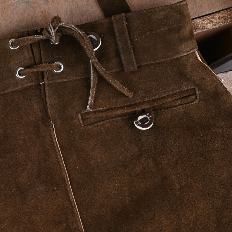 May include: A dark brown suede skirt featuring a high waistband with belt loops, a front lace-up closure with silver-toned metal grommets and matching suede laces, and a single horizontal welt pocket with a silver-toned metal button detail.