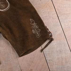May include: A dark brown suede leather short featuring white embroidered details of a deer and oak leaves. The side of the leg includes a lace-up closure with metal eyelets and a brown leather cord tied at the bottom hem.