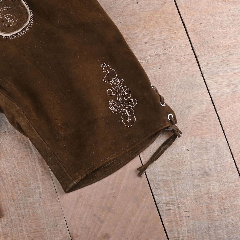 May include: A dark brown suede leather short featuring white embroidered details of a deer and oak leaves. The side of the leg includes a lace-up closure with metal eyelets and a brown leather cord tied at the bottom hem.