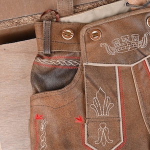May include: Brown leather lederhosen featuring a front flap with white embroidered crown and floral motifs, red decorative stitching along the edges, and two circular buttons. The garment includes belt loops and a structured waistband design.