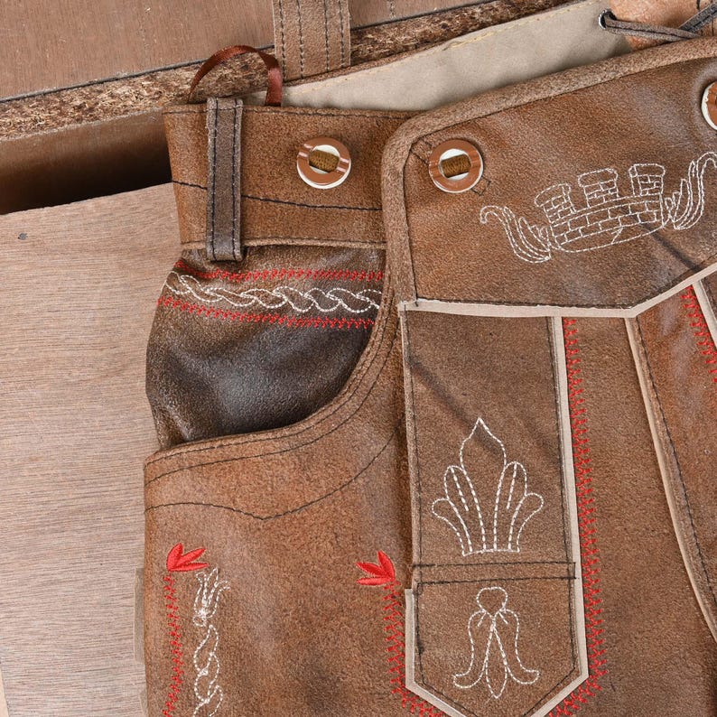 May include: Brown leather lederhosen featuring a front flap with white embroidered crown and floral motifs, red decorative stitching along the edges, and two circular buttons. The garment includes belt loops and a structured waistband design.