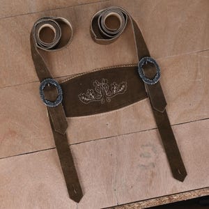May include: A dark brown suede leather suspender set for lederhosen featuring a central chest piece with light-colored decorative embroidery of oak leaves and vines, two adjustable straps with coiled ends, and two ornate silver-toned metal buckles.