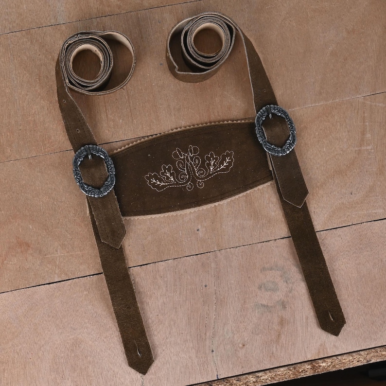 May include: A dark brown suede leather suspender set for lederhosen featuring a central chest piece with light-colored decorative embroidery of oak leaves and vines, two adjustable straps with coiled ends, and two ornate silver-toned metal buckles.