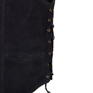 May include: Close-up of a black suede vest with a pocket and lace-up detailing. The vest has a classic design with brass-colored eyelets and a decorative stitched edge. The vest is a dark, solid color.