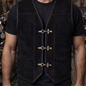 Front view of a man wearing a black suede leather biker vest with triple antique brass hook closures, styled over a black t-shirt inside a motorcycle garage. The sleeveless cowhide suede vest features braided edging along the neckline and front panels, traditional front pockets, and a structured V-cut hem. The vest is centered in the frame with blurred motorcycles in the background, highlighting rugged biker style and masculine fit.