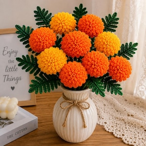May include: A bouquet of handmade crochet flowers in a white vase. The flowers are orange and yellow, with green leaves. The vase is tied with a twine bow. A framed sign reads "Enjoy the little Things".