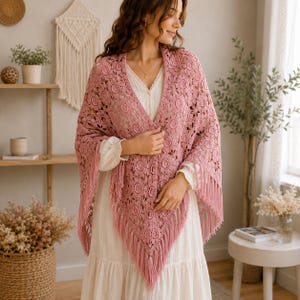 Peut inclure: Un châle triangulaire rose poudré présentant un motif en dentelle crochetée circulaire complexe et une longue frange sur les bords. Le châle est drapé sur une robe blanche à manches longues avec un devant boutonné et une jupe froncée.