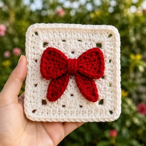Pode incluir: Um quadrado de crochê em fio branco off-white com um laço tridimensional de crochê vermelho escuro centralizado. O quadrado apresenta um padrão clássico de pontos abertos com uma borda espessa e texturizada, destacando o trabalho manual.