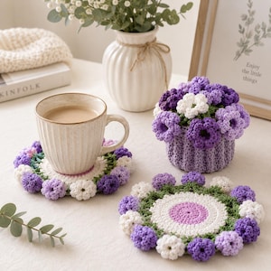 May include: A beige ceramic mug with coffee sits on a crocheted coaster with a floral design. The coaster features white, purple, and green accents. A crocheted flower basket and a vase with flowers are also in the image.