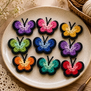 May include: Nine crocheted butterfly appliques arranged on a speckled ceramic plate. Each butterfly features a two-tone color scheme with a black crocheted border and two black antennae. The colors include purple, pink, yellow, green, blue, and red.