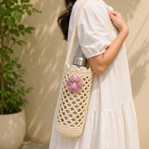 May include: A cream-colored crochet water bottle holder featuring a lattice pattern, a scalloped top edge, and a single pink flower appliqué. The cylindrical holder includes a long shoulder strap and contains a metallic silver water bottle.