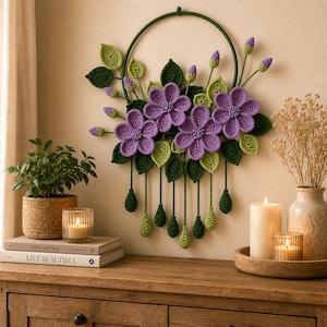 May include: A circular wall hanging featuring crocheted purple flowers and green leaves arranged on a dark green hoop. Several crocheted green leaf-shaped pendants hang from the bottom of the hoop on thin green cords, creating a decorative floral display.